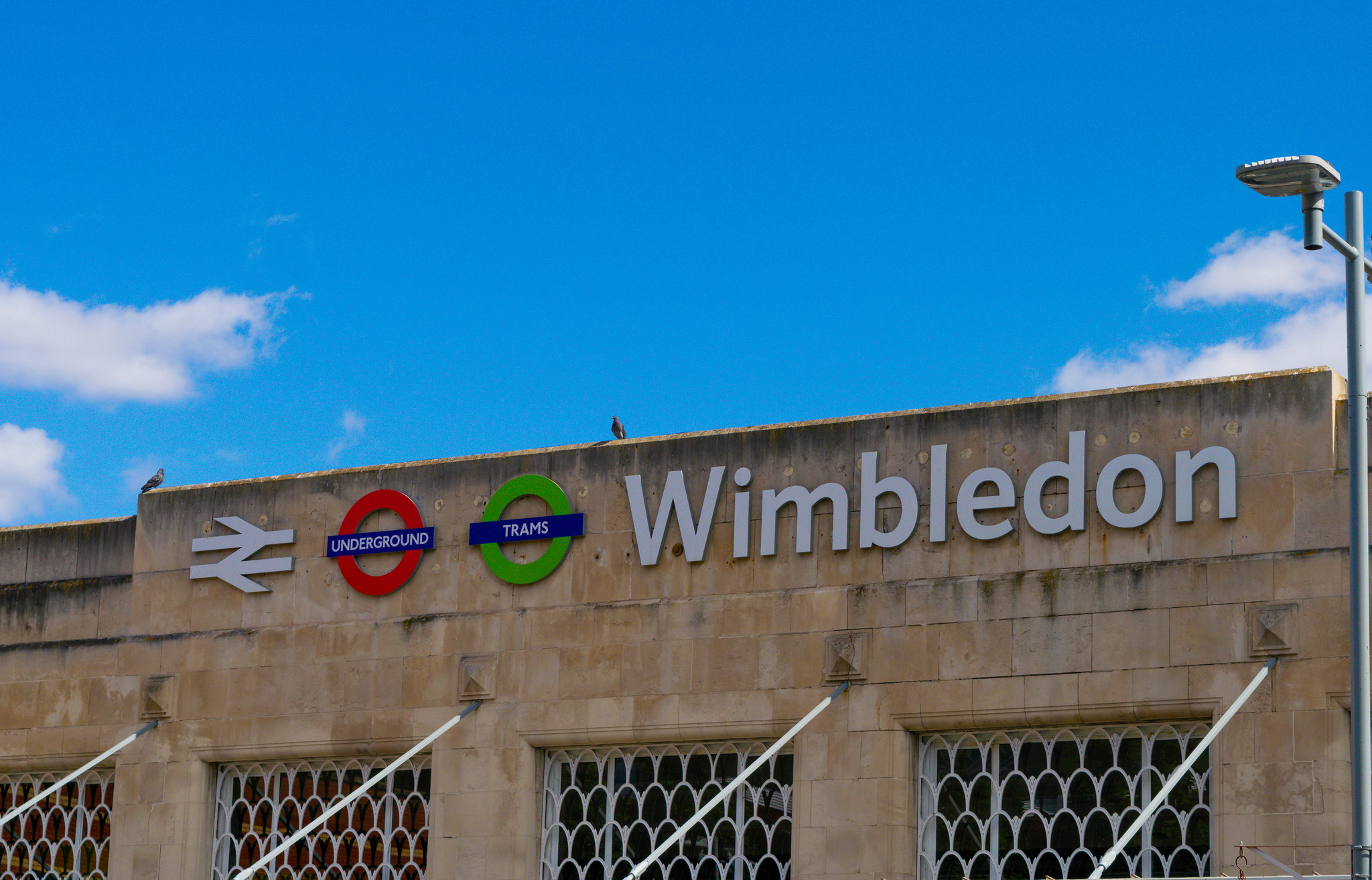 Wimbledon station sign with rail, Underground and tram symbols, marking one of the main arrival points for visitors travelling into southwest London.